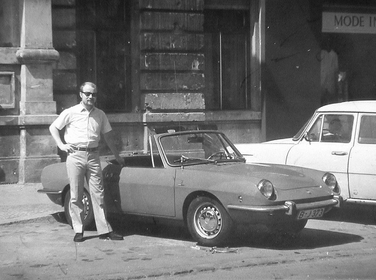 A person standing in front of a white car parked on a city street

Description automatically generated