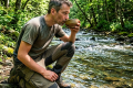 An elderly bearded man drinking from a wooden bowl by a forest stream.