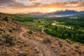 Dirt hiking trail overlooking green valley with winding river and mountain sunrise